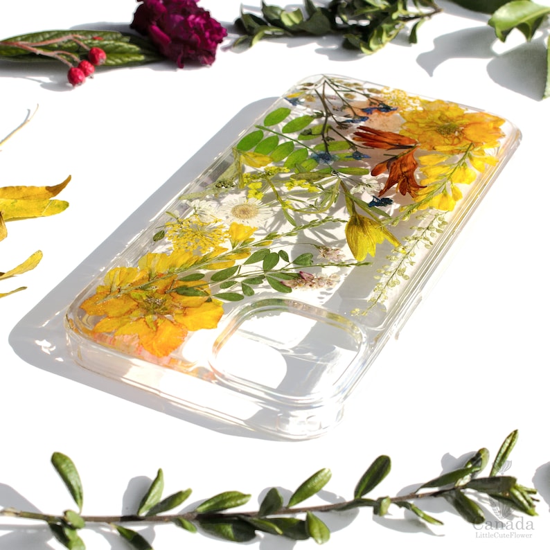 May include: A clear phone case with pressed flowers and leaves. The flowers are yellow, orange, and white. The case is designed for a smartphone with a camera cutout.