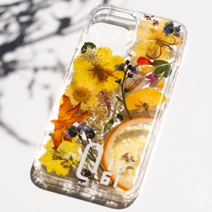 May include: A clear phone case with a pressed flower design. The case features yellow, orange, and white flowers, as well as a slice of lemon. The case has a white logo that says "0161".