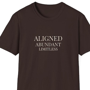 Puede incluir: Una camiseta marrón oscuro con las palabras "ALIGNED, ABUNDANT, LIMITLESS" impresas en color beige claro. La camiseta tiene cuello redondo y mangas cortas. El texto está centrado en la parte delantera.