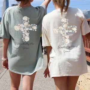 May include: Two people wearing light green and cream colored t-shirts with a floral cross design on the back. The shirts have the text "Washed by His Grace" printed on the back.