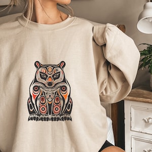 May include: Beige sweatshirt with a detailed bear design in black, orange, and red. The bear graphic has intricate patterns and a stylized appearance. The sweatshirt is a neutral color, suitable for casual wear.