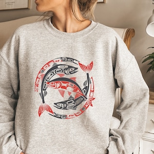 May include: A gray sweatshirt with a stylized design of three salmon swimming in a circle. The salmon are black, red, and white.
