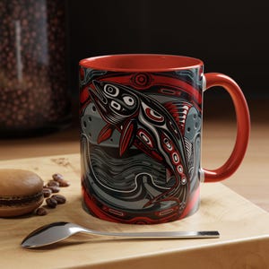 May include: A red-handled coffee mug with a Native American-inspired design. The mug features a stylized salmon illustration in black, red, and white against a gray background. The interior of the mug is red.