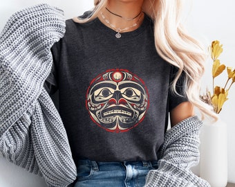May include: A dark gray t-shirt with a stylized graphic of a Native American face in red, white, and black colors. The graphic is circular and has a red border.