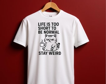 May include: White t-shirt featuring a black illustration of a grumpy cat with a coffee cup, and the slogan "LIFE IS TOO SHORT TO BE NORMAL STAY WEIRD". The tee is suspended from a wooden hanger.