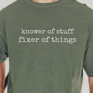 May include: Olive green t-shirt with the text "knower of stuff fixer of things" in white. The shirt is a relaxed fit and has short sleeves. The person is wearing blue jeans and a brown watch.