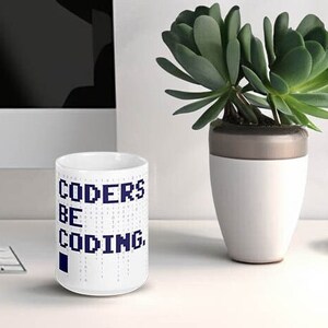 Puede incluir: Taza de café de cerámica blanca con un diseño azul que dice "CODERS BE CODING". La taza está sobre una mesa blanca.