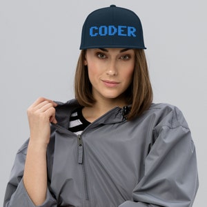 Può includere: Un cappello da baseball blu navy con testo pixelato blu che recita "CODER" sul davanti.