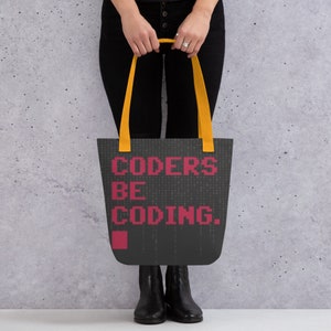 Op de afbeelding: Een zwarte tote bag met gele handvatten en de tekst "CODERS BE CODING." in roze gepixelde letters. De tas heeft een zwart-wit gepixelde achtergrond.