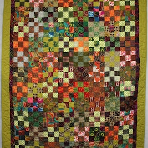 May include: A rectangular quilt featuring a patchwork grid of small squares in various colors including green, orange, red, and brown. The quilt has a dark patterned inner border and a wide solid olive green outer border with quilted stitching.