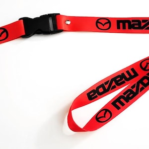 Puede incluir: Cordón rojo con el logotipo y texto Mazda en negro. El cordón tiene una hebilla de plástico negro y un cierre de metal plateado. El logotipo de Mazda es un círculo estilizado con una línea curva.