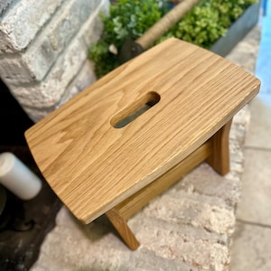 May include: A wooden stool with a rectangular top and a cutout handle. The stool is made of light brown wood and has a natural finish.