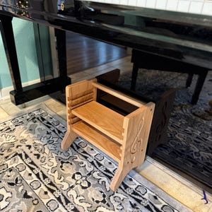 May include: A wooden step stool with two shelves and a treble clef design. The stool is made of light brown wood and has a natural finish. The stool is sitting on a patterned rug in front of a black piano.