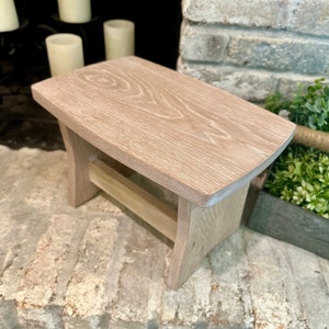 May include: A whitewashed wooden stool with a simple design. The stool has a rectangular top and a single shelf underneath. The stool is made of natural wood and has a rustic look.