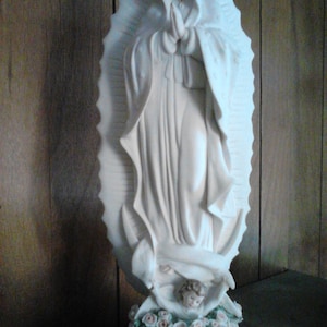 May include: White porcelain figurine of the Virgin Mary, standing with hands clasped in prayer. She is wearing a flowing robe and a crown. The figurine is surrounded by white roses and an angel at her feet.