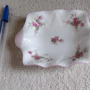 May include: A small, white, porcelain dish with a scalloped edge and pink accents. The dish is decorated with pink roses and green leaves. A blue pen is next to the dish.
