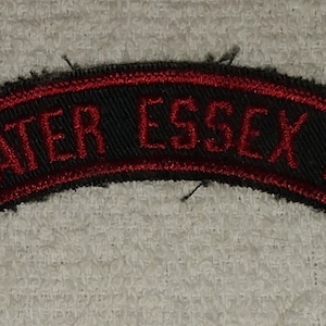 May include: A black and red embroidered patch with the text "GREATER ESSEX N.J." in red. The patch has a curved shape and is likely a collectible or souvenir item.