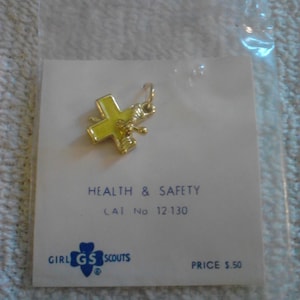 May include: A gold-colored cross-shaped charm with yellow enamel, attached to a small card. The card reads "HEALTH & SAFETY" and "GIRL SCOUTS". The charm is in a clear plastic bag.