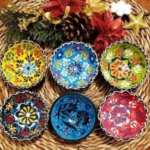May include: Six small, colourful ceramic bowls with intricate floral patterns. The bowls are arranged in a circle on a woven straw surface.
