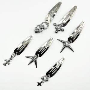 Peut inclure: Six pinces à cheveux en métal argenté avec divers breloques pendantes, dont une croix, une étoile et des pointes. Les pinces sont disposées sur une surface blanche, mettant en valeur leur design élégant et moderne. Accessoires pour un style audacieux.