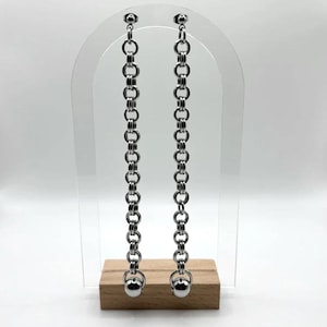 Puede incluir: Un par de pendientes largos de cadena plateada. Cada pendiente presenta una serie de eslabones circulares conectados, que culminan en una pequeña bola plateada. Los pendientes se exhiben sobre un soporte transparente con base de madera.