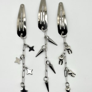 May include: Three silver hair clips with dangling chains. One chain has star charms, another has cone-shaped spikes, and the third has tooth charms. The clips are on a white background.