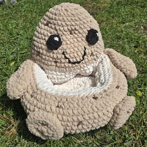 May include: A beige crochet plush toy shaped like a cookie with a hollow centre. The top features a smiling face with black eyes and a stitched mouth. The exterior is textured with small brown spots, while the interior is lined with white yarn.