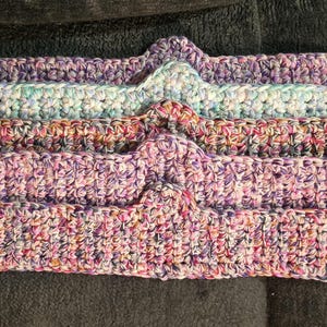 May include: A stack of five handmade crochet headbands in various colors. The headbands are primarily purple, pink, and blue, with a textured, woven appearance. Each headband has a decorative, pointed top.
