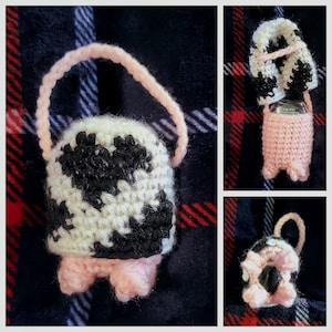 May include: A crocheted cow-print purse with a pink handle and pink feet. The purse is made of black and white yarn and has a black and white cow print design.