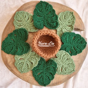 May include: A set of eight crocheted monstera leaf coasters in shades of green. The coasters are arranged in a circle around a small brown crocheted basket with the text "Yarn On" in white.