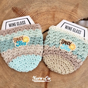 Peut inclure: Deux chauffe-verres en crochet de couleur beige, bleu clair et vert avec un patch brodé "Sandy Toes" sur chacun. Les chauffe-verres sont posés sur une surface en bois.