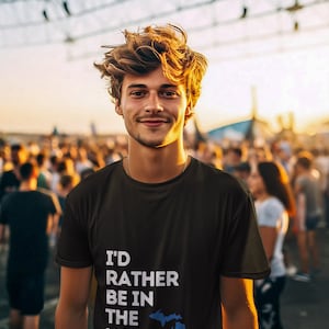 Può includere: Un uomo indossa una maglietta nera con testo bianco che recita "I'd rather be in the U.P." e un contorno blu dello stato del Michigan. È in piedi di fronte a una folla di persone.