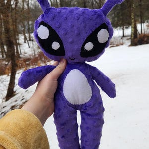 May include: A plush purple alien doll with large black and white eyes, two antennae, and a white belly patch. The doll is made of soft, textured fabric and has a smiling face. The background is a snowy outdoor scene.