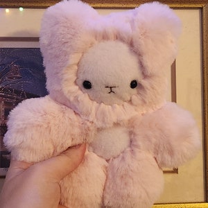 Könnte beinhalten: Ein weicher, rosa Plüschhase mit weißem Fell an Gesicht und Bauch. Der Hase hat schwarze Augen und eine kleine schwarze Nase.