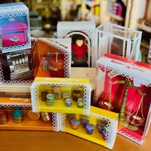May include: A collection of Bodo Hennig miniature toy sets in clear plastic boxes with white lace-patterned borders, featuring a red mailbox, a silver cash register, a brass balance scale, and various small food containers and jars.