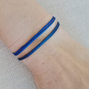 May include: Two blue cord bracelets on a person's wrist. The bracelets are made of a thin, braided cord and are tied in a simple knot.