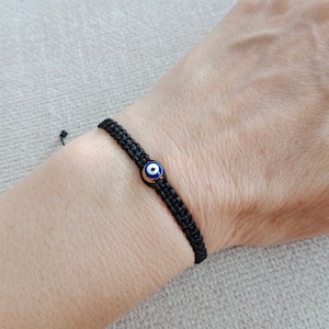 May include: A black string bracelet with a blue and white evil eye charm. The charm is gold-toned.