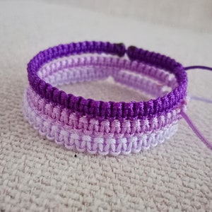 May include: A purple, lavender, and white ombre friendship bracelet made with braided cord.