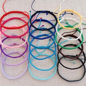 May include: A collection of adjustable braided cord bracelets in various colors including red, pink, purple, navy blue, light blue, yellow, white, green, brown, and black. Each bracelet features a woven pattern with loose pull strings for sizing.