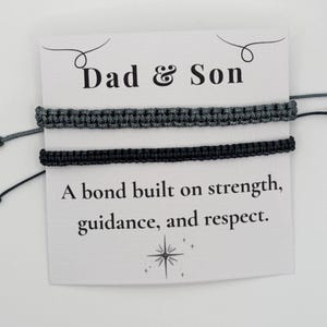 May include: Two macrame bracelets, one gray and one black, are displayed on a white card. The card reads "Dad & Son" above the bracelets and "A bond built on strength, guidance, and respect" below.