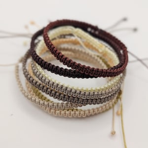 May include: A stack of five adjustable cord bracelets featuring a woven macrame pattern. The bracelets are colored in shades of dark brown, cream, light gray, taupe, and beige, each with thin trailing cords extending from the sides for closure.