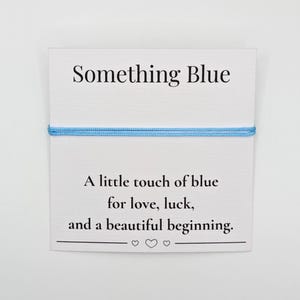 May include: A white card with the text "Something Blue" and a light blue string bracelet. The card also reads "A little touch of blue for love, luck, and a beautiful beginning."