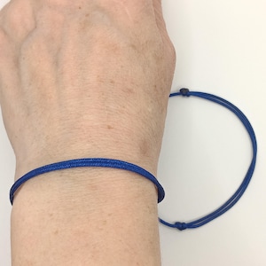 May include: A blue cord bracelet with a simple knot closure. The bracelet is shown on a person's wrist.