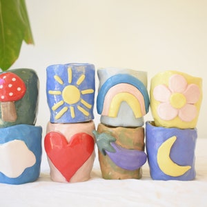 May include: A selection of eight small, handmade ceramic pots. Each pot is a different colour and decorated with whimsical designs, including a mushroom, sun, rainbow, flower, cloud, heart, aubergine, and moon. These decorative pots are ideal for small plants.
