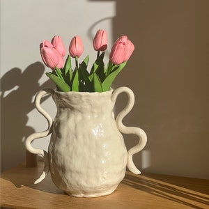 Puede incluir: Un jarrón de cerámica blanca con dos asas en forma de bucle. El jarrón está lleno de tulipanes rosas.