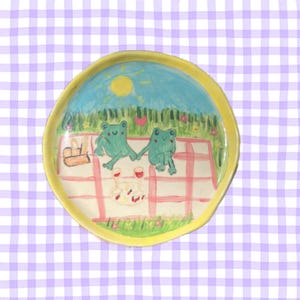 Può includere: Un piatto in ceramica dipinto a mano con due rane verdi su una tovaglia da picnic a quadri rossi e bianchi. La scena include un sole, erba e fiori.