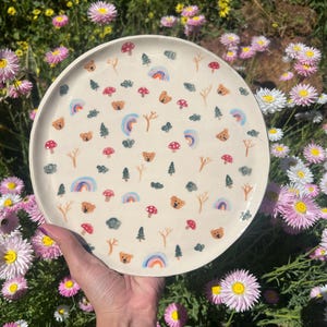 Pode incluir: Um prato de cerâmica creme decorado com um padrão lúdico de arco-íris, ursinhos de pelúcia, cogumelos e árvores. O prato é segurado em frente a um fundo de flores coloridas.