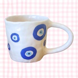 Può includere: Tazza in ceramica bianca con manico arrotondato, decorata con disegni blu e bianchi a forma di occhio. La tazza ha una superficie strutturata e un design semplice ed elegante, perfetta per caffè o tè.