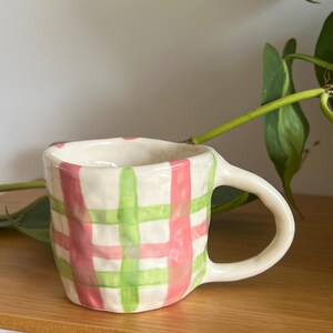 May include: A handmade ceramic mug with a cream-colored base, adorned with a pink and green plaid pattern. The mug has a rounded handle and is displayed on a wooden shelf. The design is reminiscent of a picnic blanket.