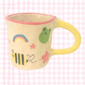 May include: A cream-coloured ceramic mug with a yellow handle and a pink rim. The mug is decorated with a rainbow, a bee, a frog, a flower, and a heart. A delightful and unique mug for tea or coffee, perfect for any kitchen.